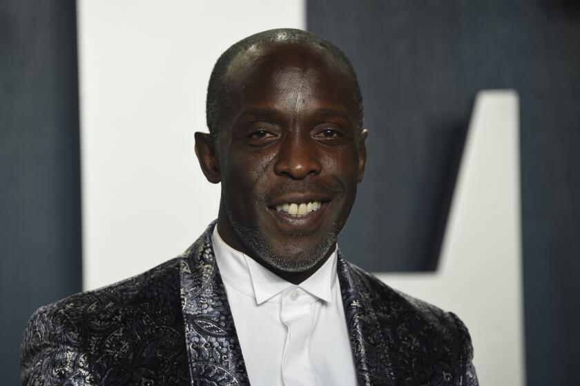 Michael K. Williams in a silver suit jacket and a dress shirt smiling in front of a grey background