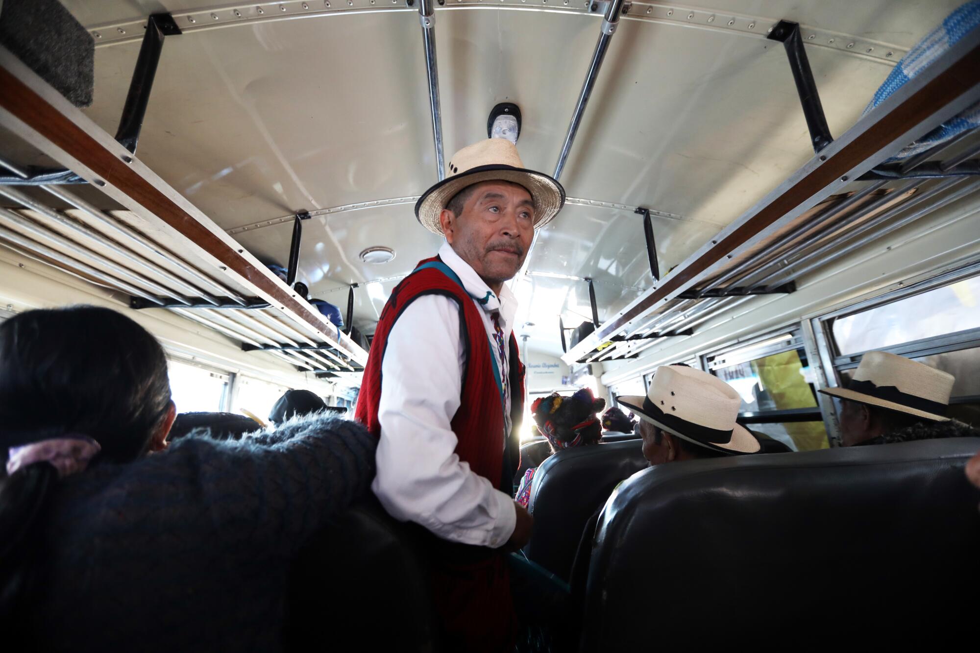 A man in a hat, white long-sleeved shirt and red vest stands near passengers in a bus 