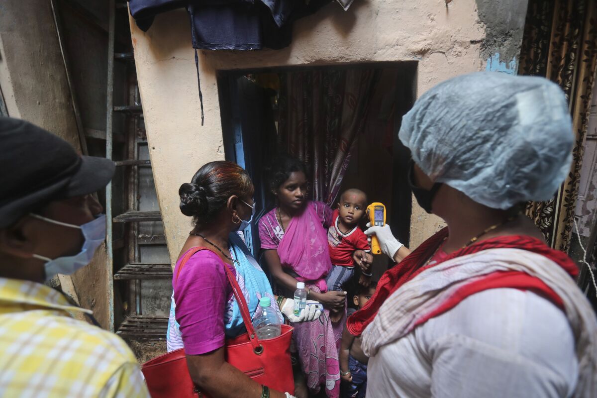 A woman in a sari holds her child within a doorway as another woman takes his temperature with a forehead thermometer.
