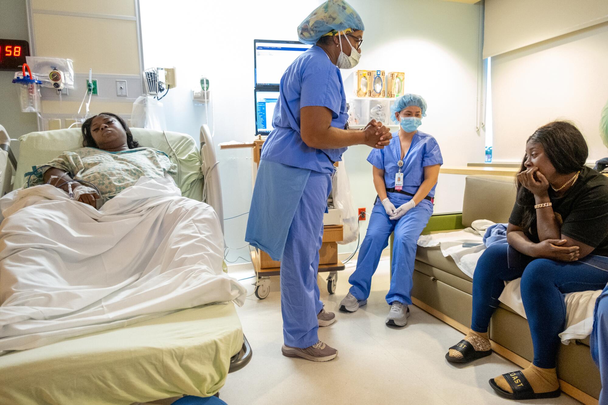 Two women in nursing scrubs talk with a seated woman as another woman lies in bed.