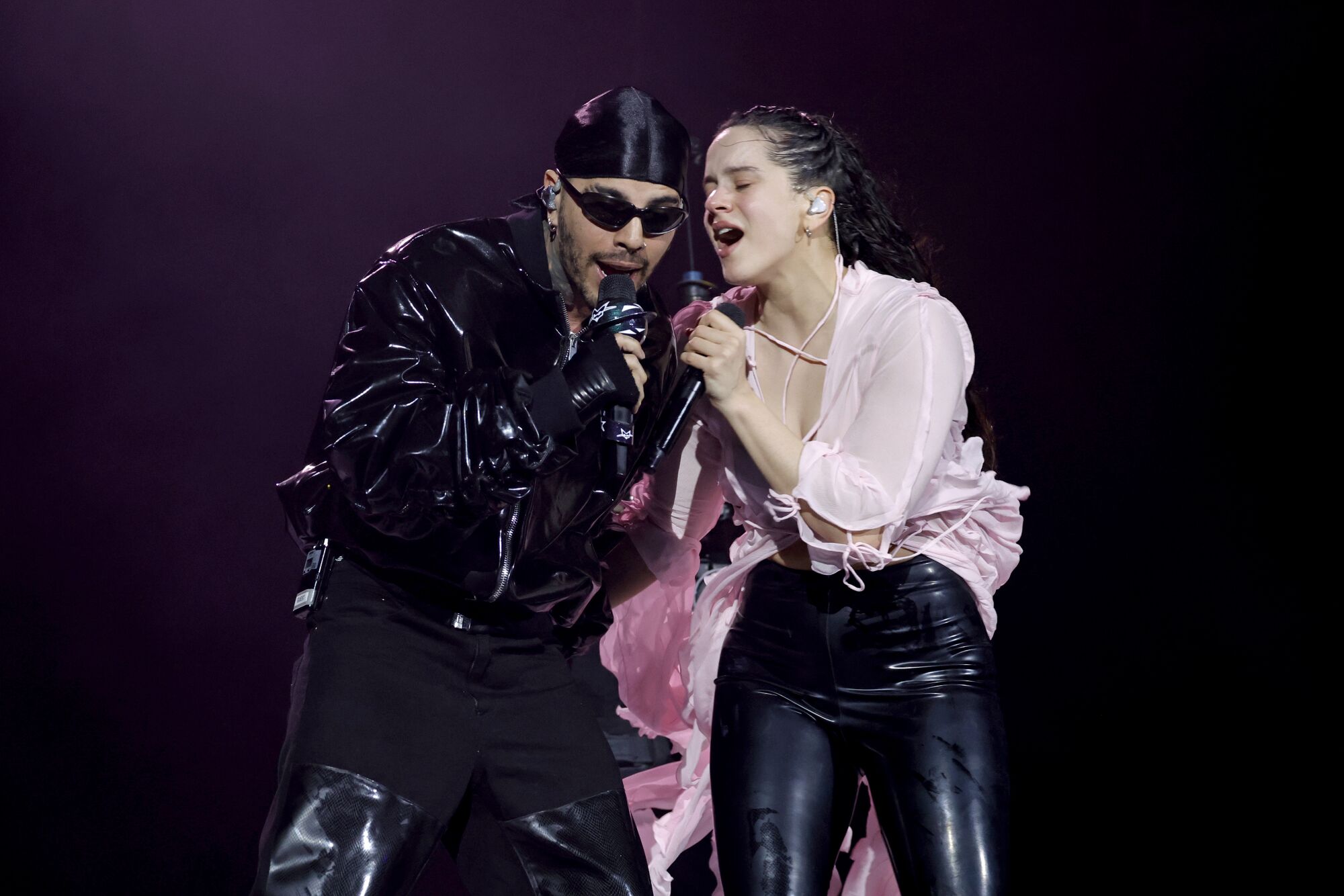 A man in black and a woman in a pink top singing into a handheld microphone on stage