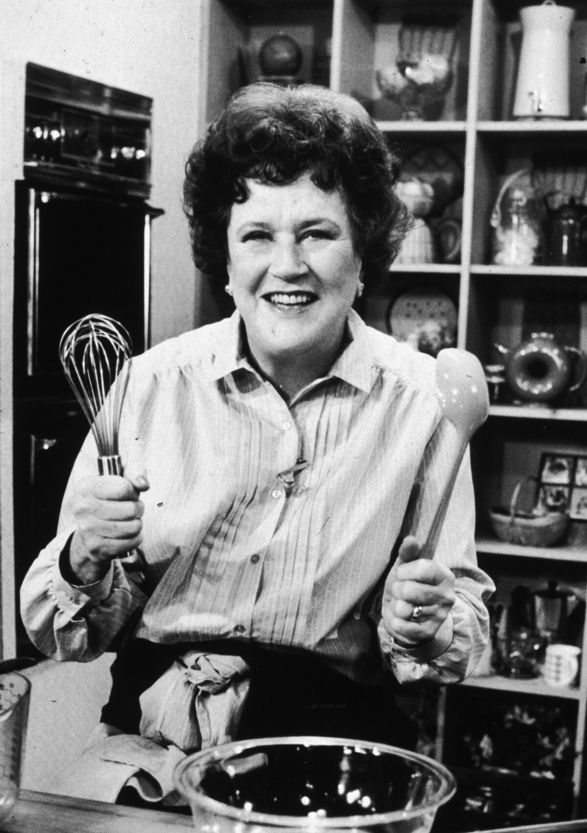 Black-and-white photo of chef Julia Child standing in front of a countertop, holding a whisk and a ladle by a mixing bowl