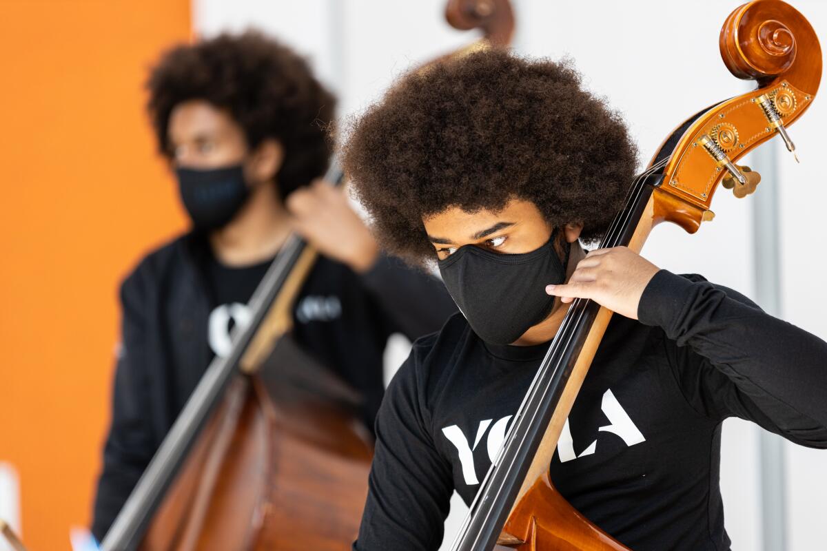 Two young men, wearing masks, stand as they play cellos.