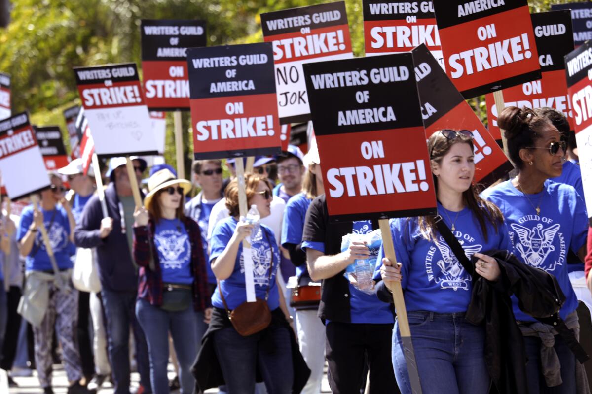 a crowd of people in a picket line with black and red signs reading "Writers Guild of America on Strike!"