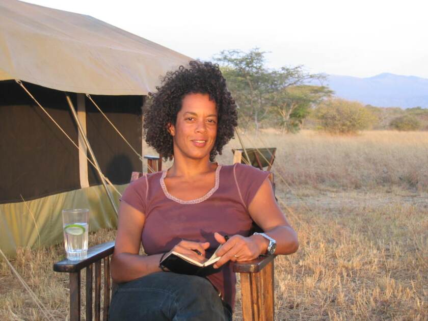 A woman sits in a chair, with a glass of water sitting on the arm, near a tent while taking notes in a book.