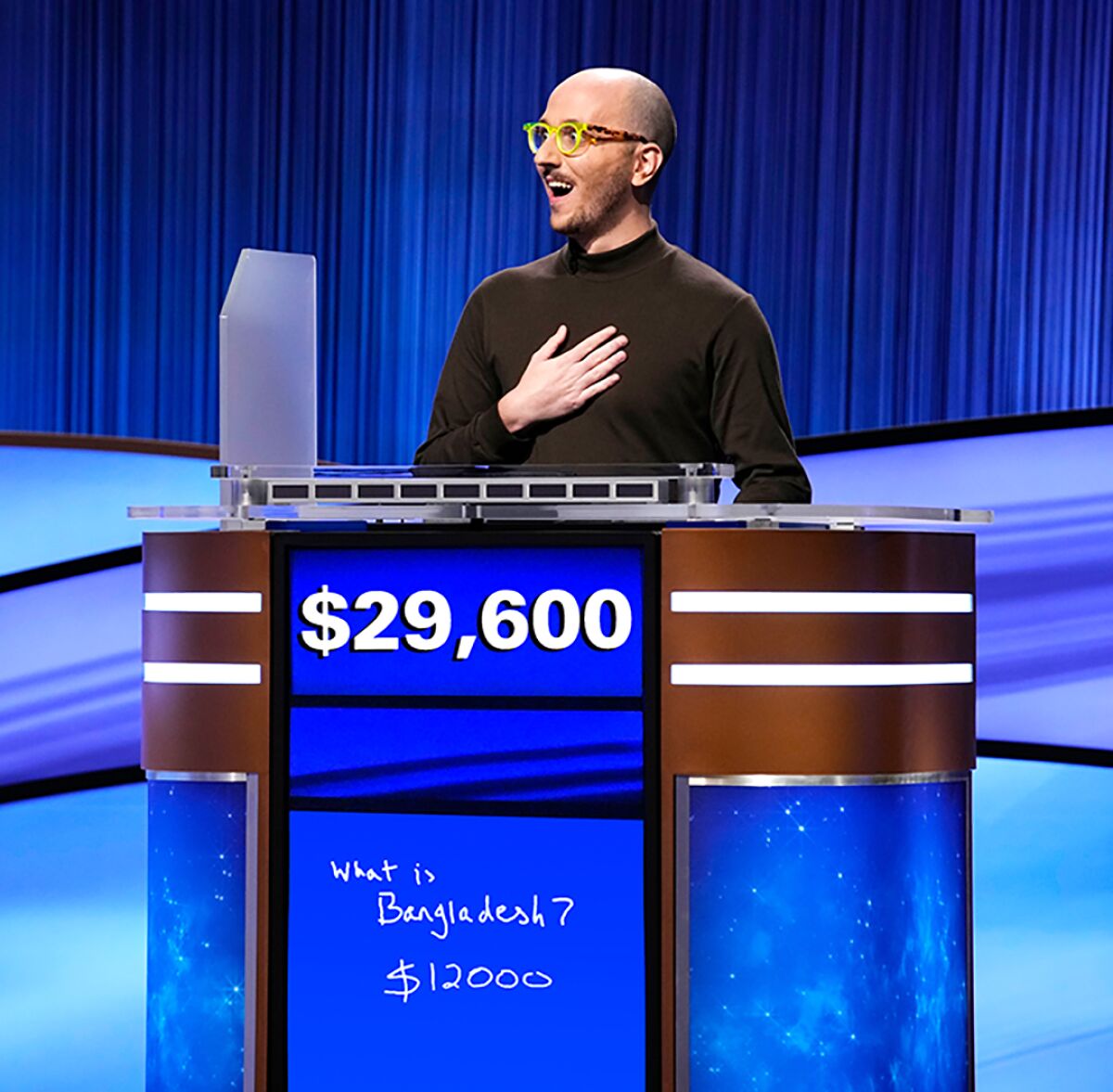 A man in a brown turtleneck holds his hand over his chest behind a "Jeopardy!" podium.