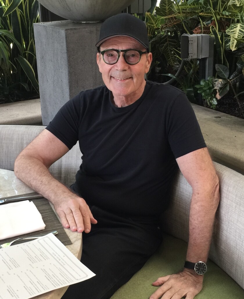 مردی با تی شرت مشکی، عینک لبه دار مشکی و کلاه بیسبال سیاه در یک غرفه رستوران نشسته است.