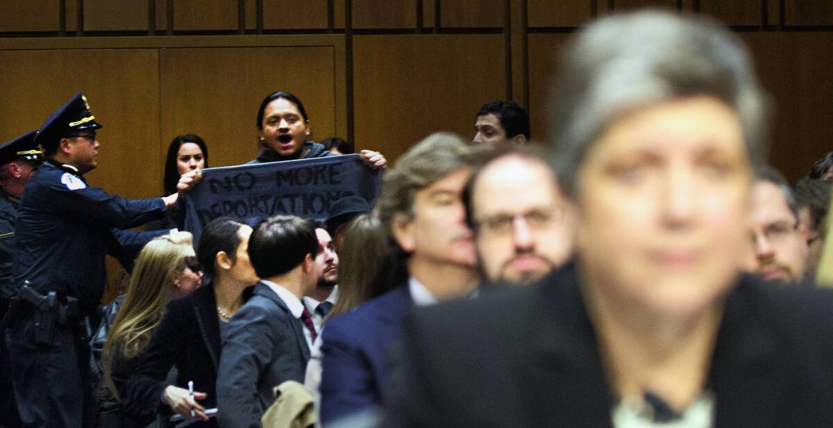A protester holds a banner that reads "No More Deportations" at a Senate committee hearing featuring Homeland Security Secretary Janet Napolitano, foreground.