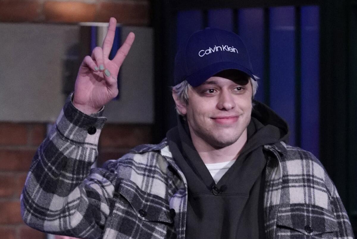 Pete Davidson, wearing a plaid shirt-jacket, hoodie and blue Calvin Klein cap, flashes a peace sign.