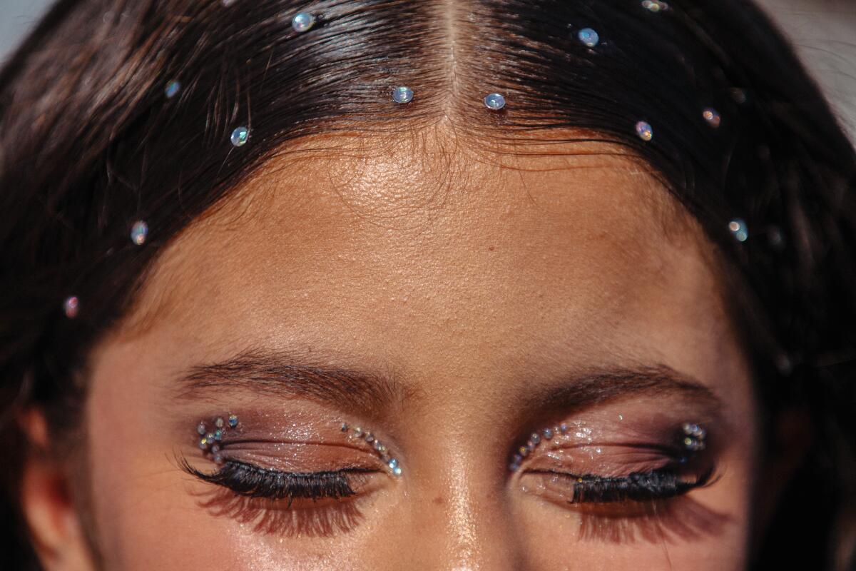 A close-up of a young woman's sparkly eyelids, and gems on her hair.