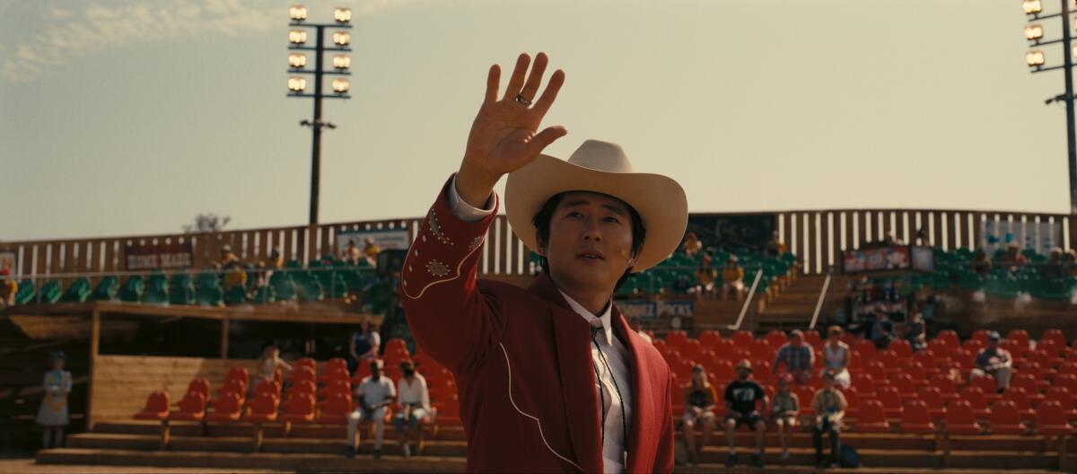 Steven Yeun in a cowboy suit waves at people seated at an outdoor theme park attraction