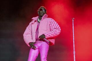 Young Thug wearing an oversized pink puffer jacket and tight hot pink pants on a stage. He stands by a mic stand