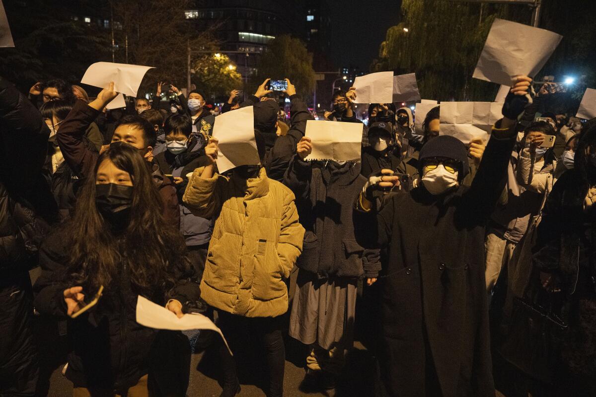 A crowd of people in warm clothing at night holding up blank papers, many in front of their faces.