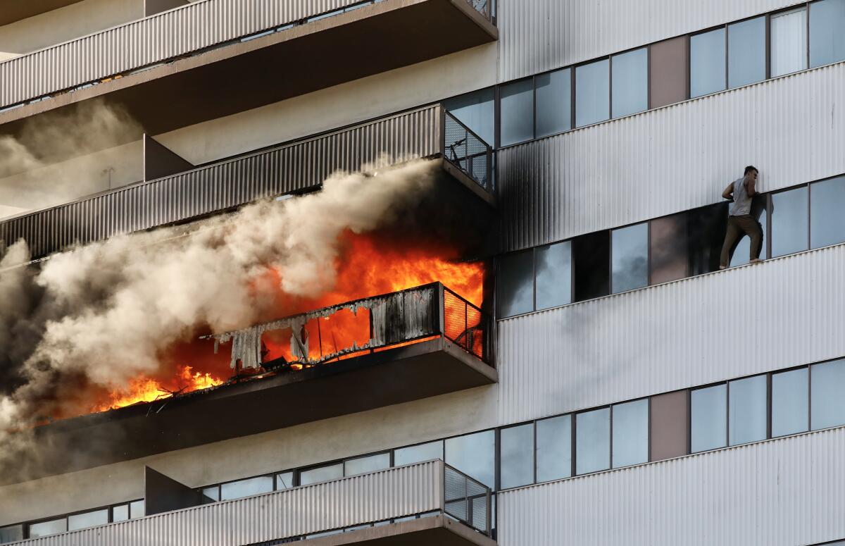 A resident holds on standing on a ledge outside an apartment window while a nearby balcony is engulfed in flames. 