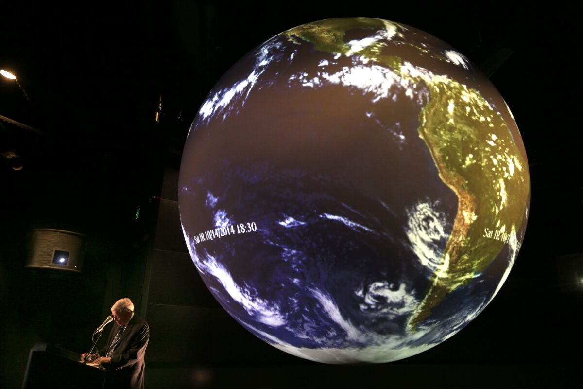A man in a dark room stands in front of a massive digital globe showing weather patterns on Earth.