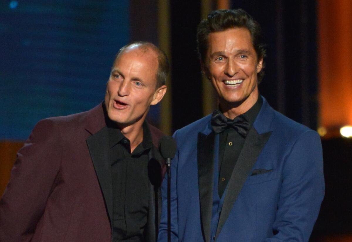 Woody Harrelson wearing a maroon suit and Matthew McConaughey wearing a navy blue suit smiling on stage.