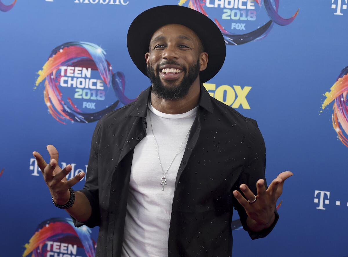 A man wearing a hat smiles and holds his hands out while standing in front of a backdrop.