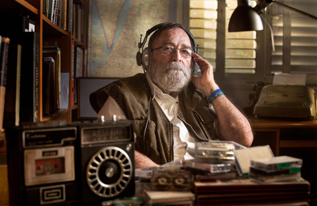 A man with big headphones on at a desk with piles of cassettes 