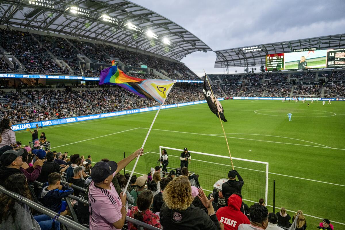 A person standing in soccer stadium seating waves a Pride flag.