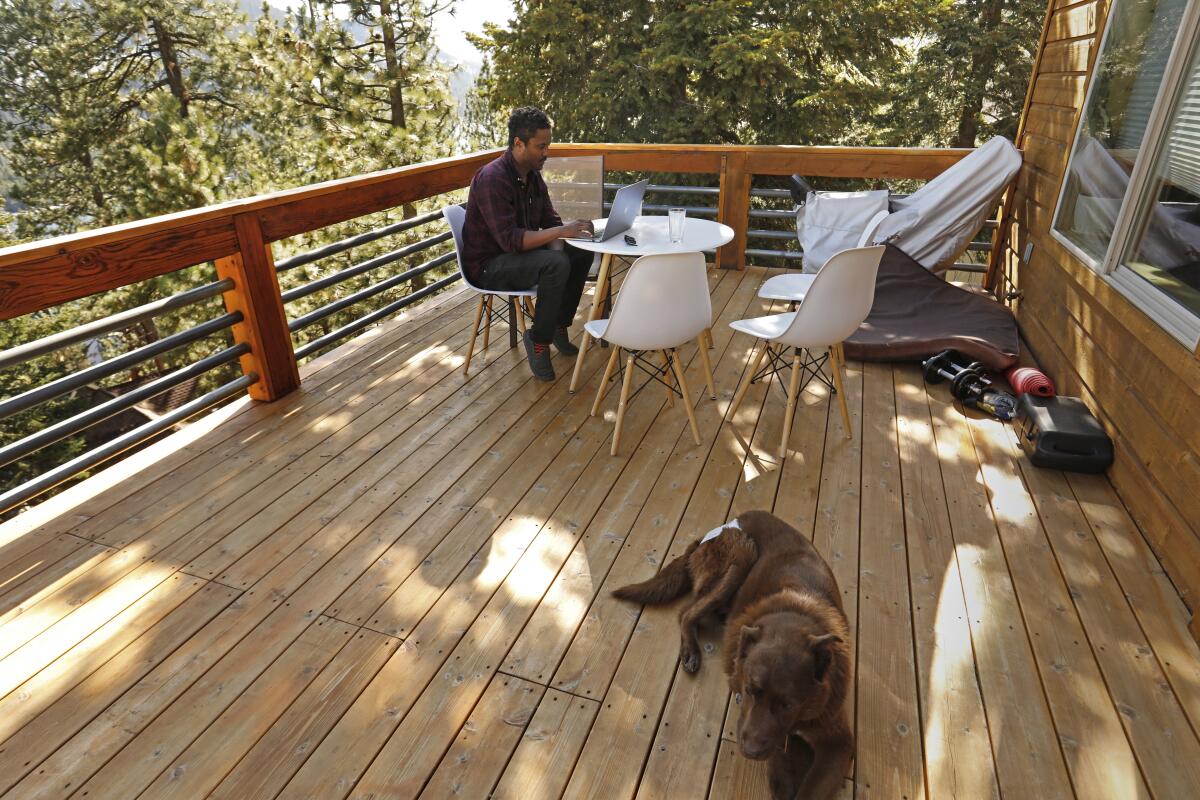 A man works on a laptop at a table outdoors on a deck while a dog sits nearby