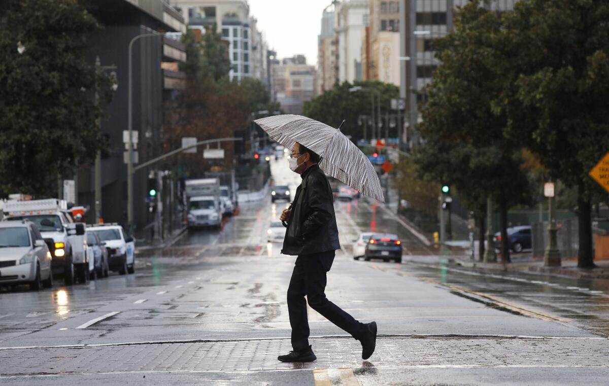A man wearing a face mask holds an umbrella as he crosses a street in downtown Los Angeles.