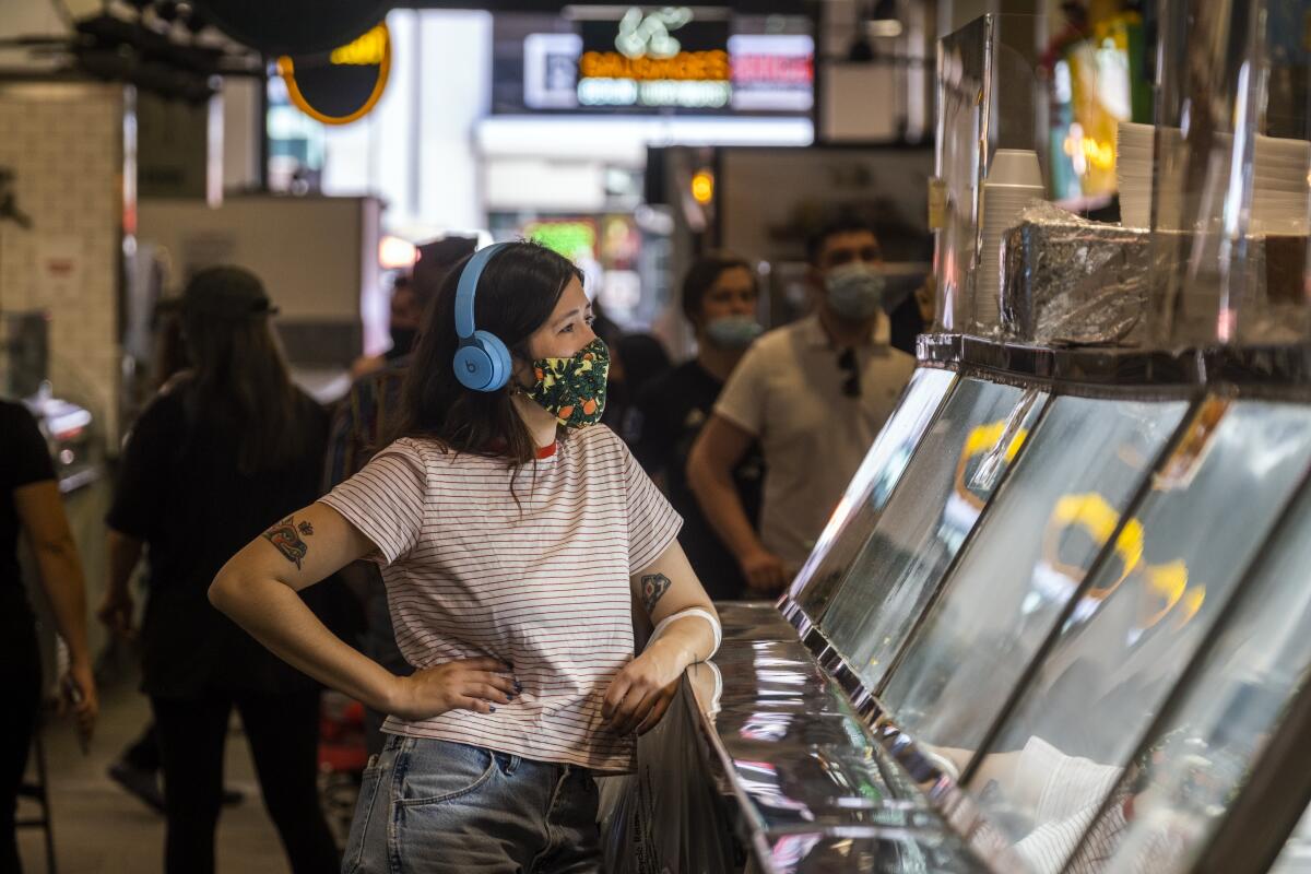 A woman in a face mask and headphones leans one arm on a deli counter.