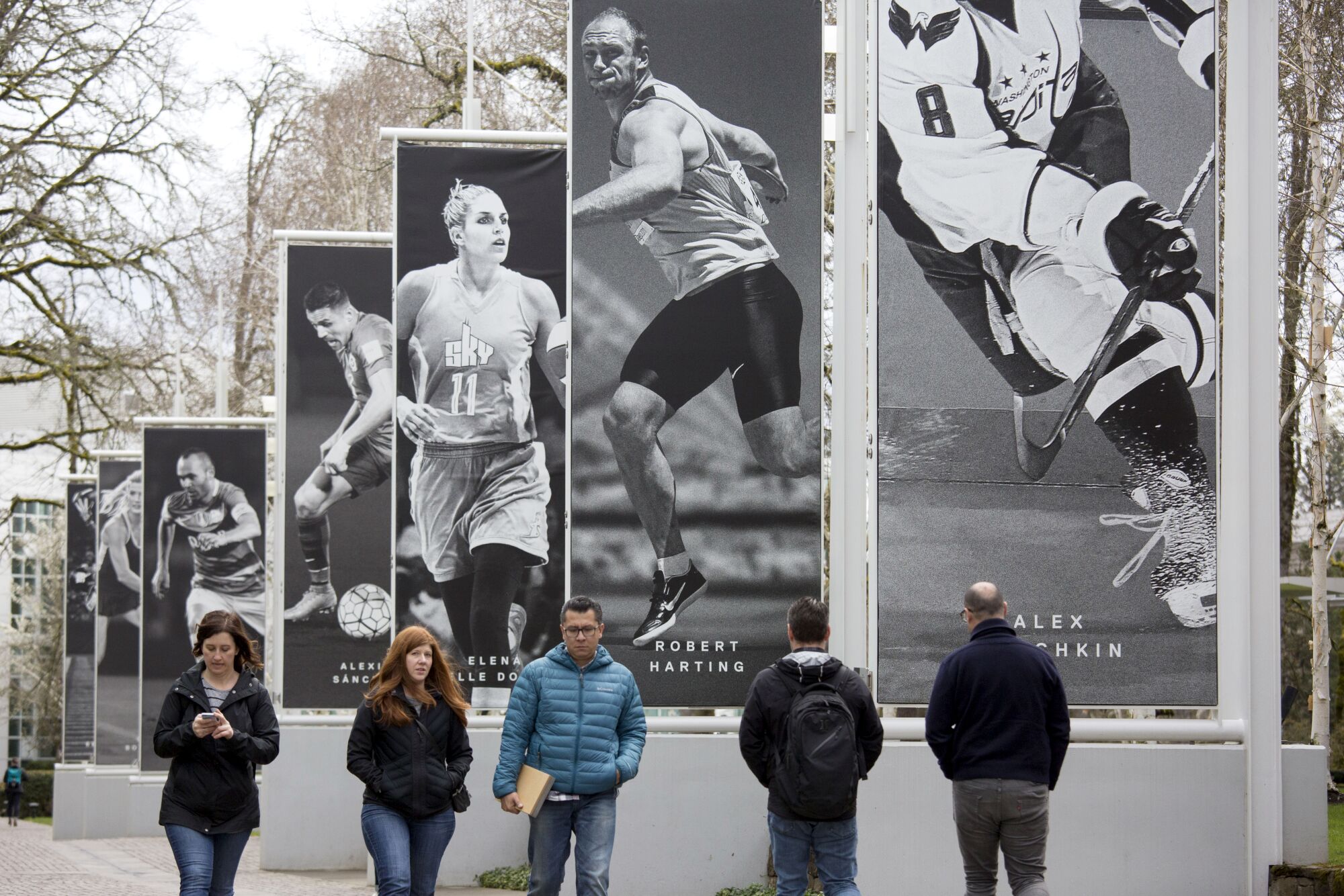 Plusieurs personnes marchant sur un trottoir à côté d'une série de bannières en noir et blanc représentant des athlètes.