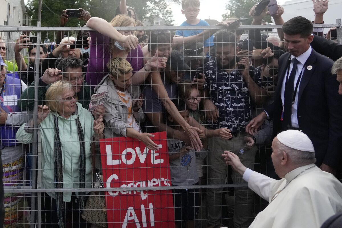 A man in a white skullcap and vestments reaches a hand out to crowds extending their hands to him behind a fence 