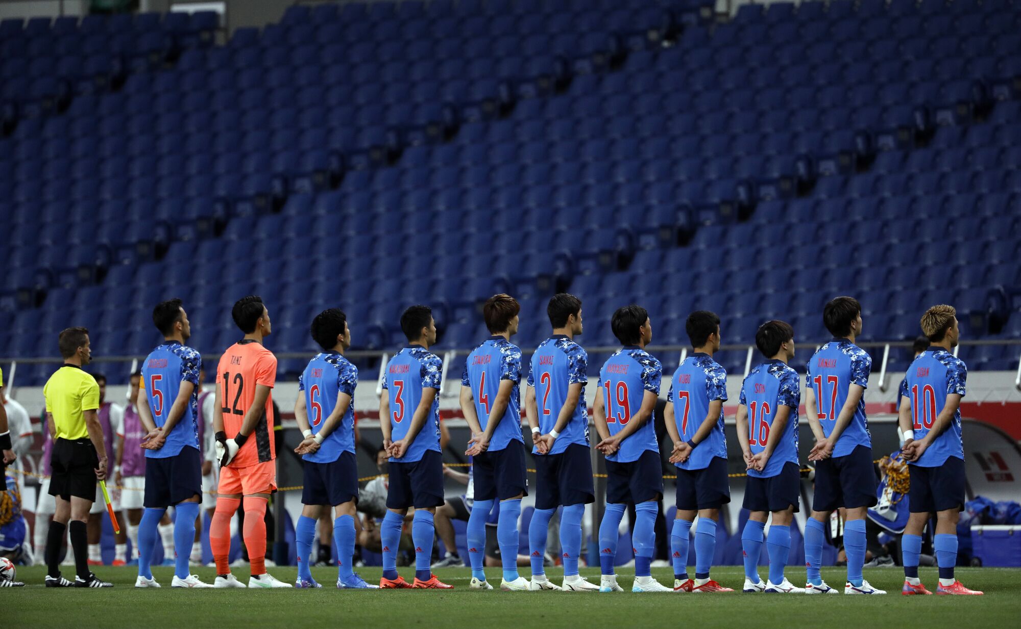 Soccer players stand in a line, hands clasped behind their backs, facing rows of empty stadium seats.