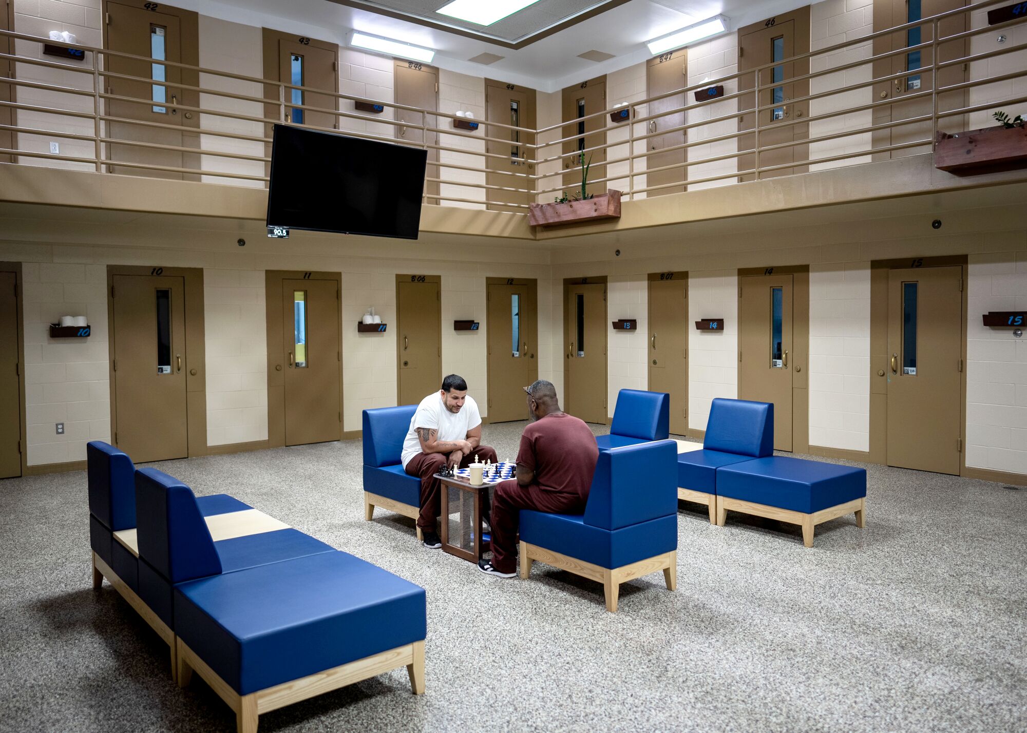 Two inmates sit in the center of a prison block and play chess.  A television is mounted on the railing of the second floor of the cellular units.