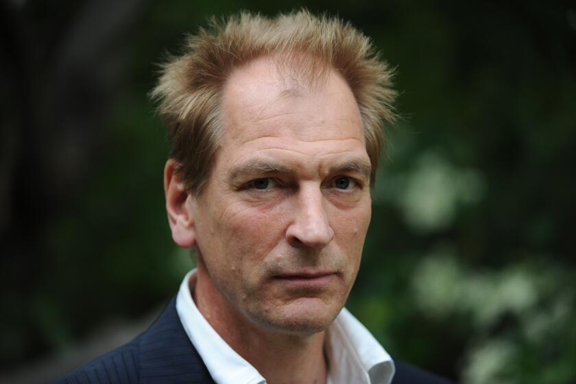 Julian Sands in a black blazer and white suit shirt against a blurry green background