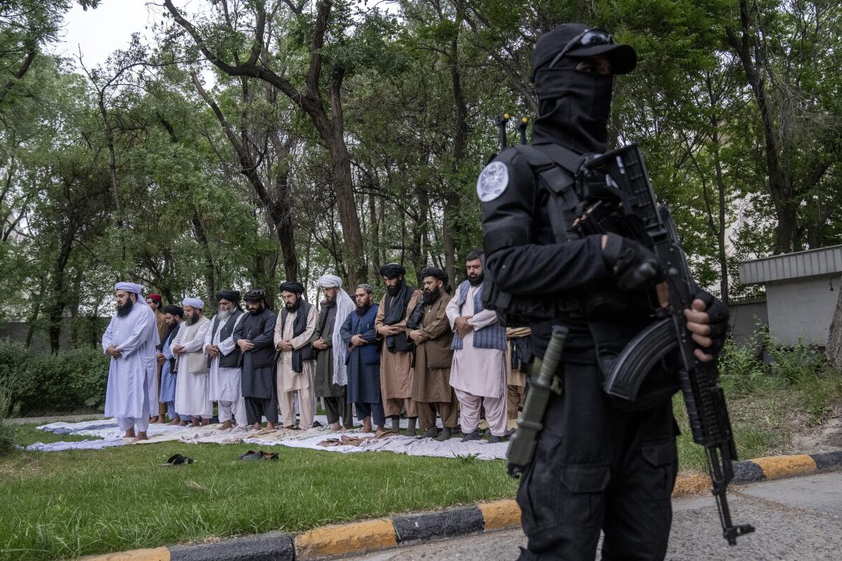 An armed man in black guards the scene in front of a line of Taliban men in traditional dress