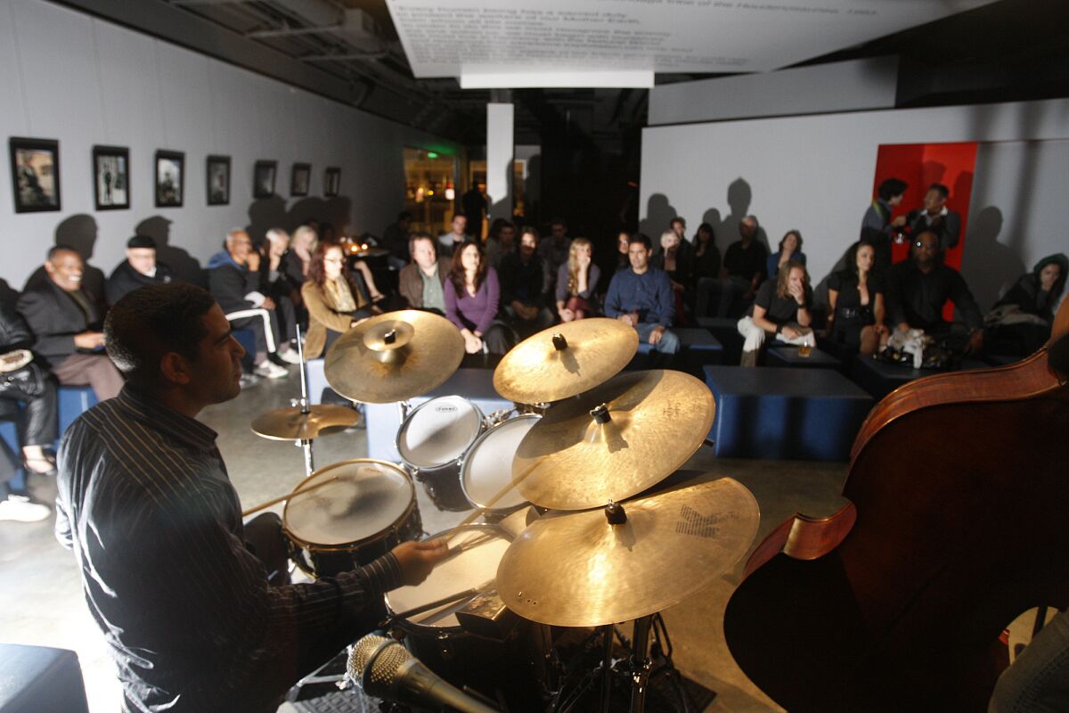 A drummer sits at his set in front of a small crowd seated a dim jazz club