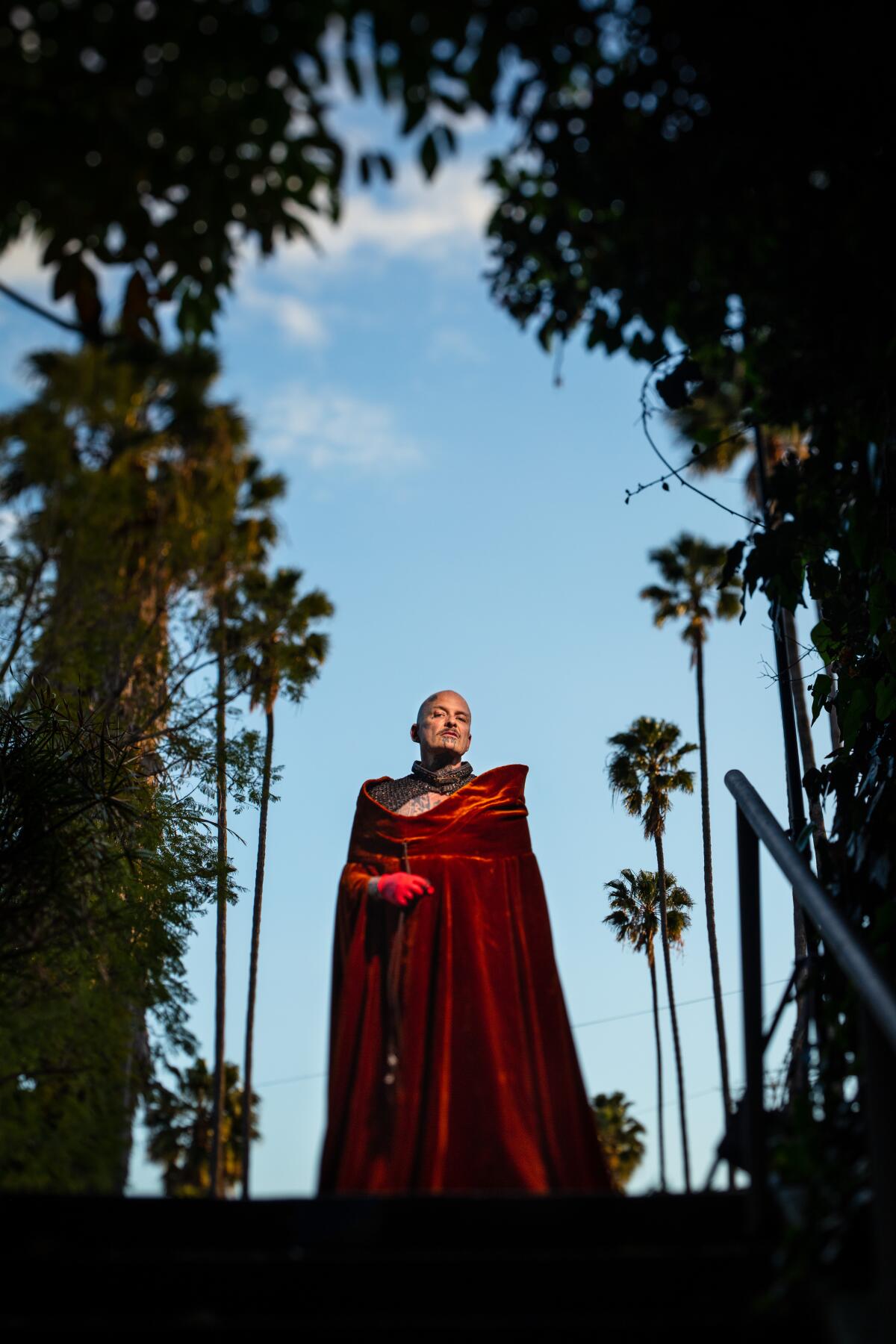 Performance artist Ron Athey, wearing a red cape, stands among trees.