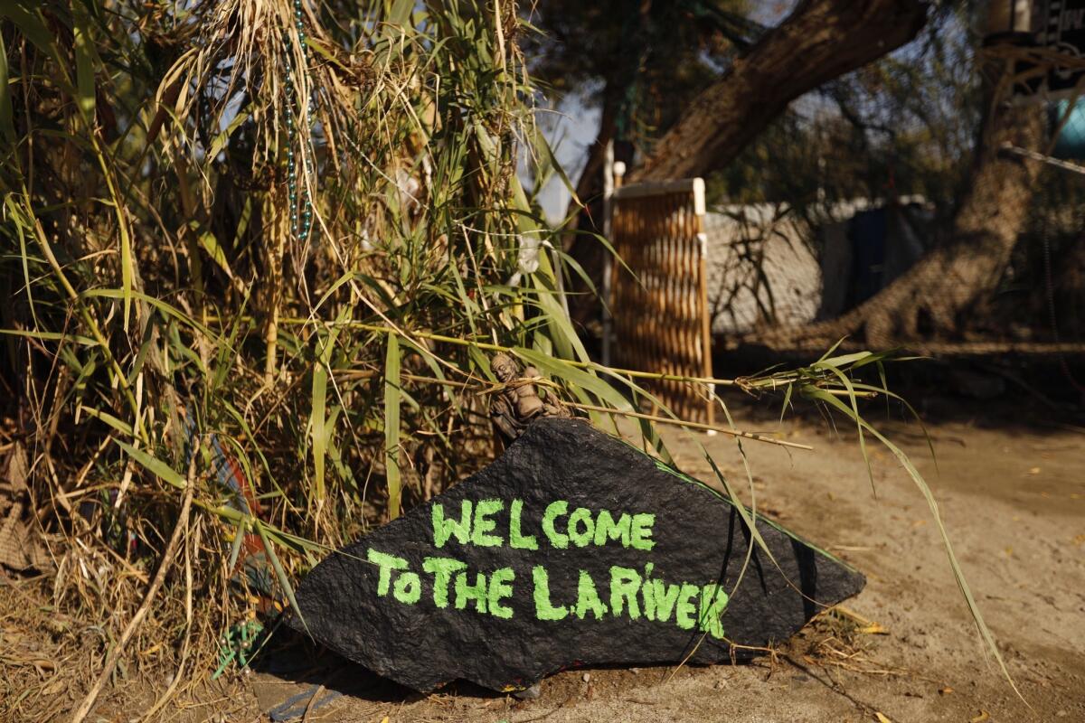 "Welcome to the L.A. River," reads a painted rock that greets visitors at the entrance to a campsite shared by Melissa Millner, 46; Justin Reeves, 46; and Tyrone Hart, 56. They live on an island in the middle of the river. The river's earthen center is thick with reeds and trees.