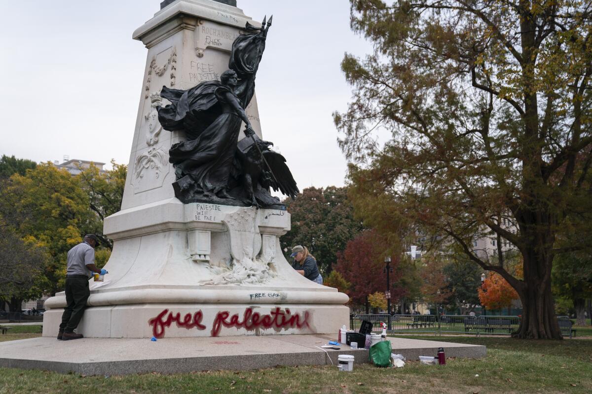 Workers clean a vandalized sculpture in a park; visible is graffito reading "Free Palestine."