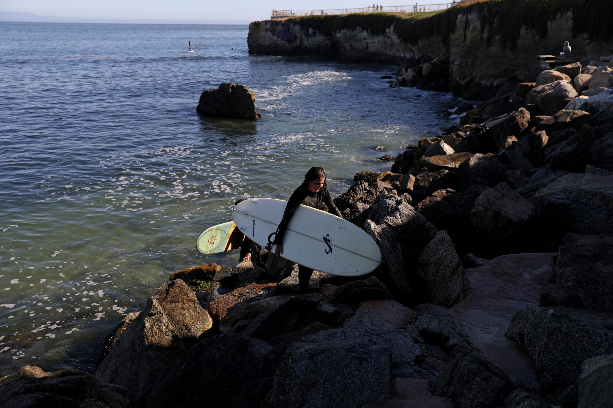 A surfer holding a white board walks up a rocky incline 