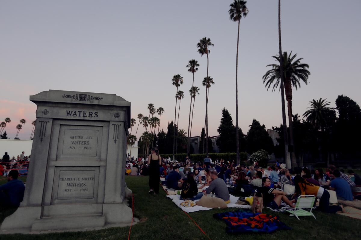 People sitting on blankets under palm trees at dusk, next to a large stone memorial, in a cemetery