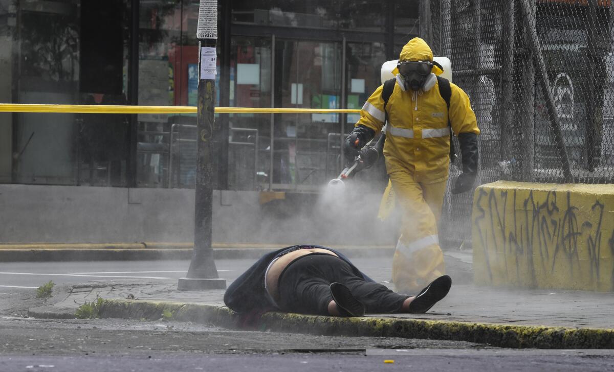 A worker in full hazmat gear sprays disinfectant on a body lying on the sidewalk