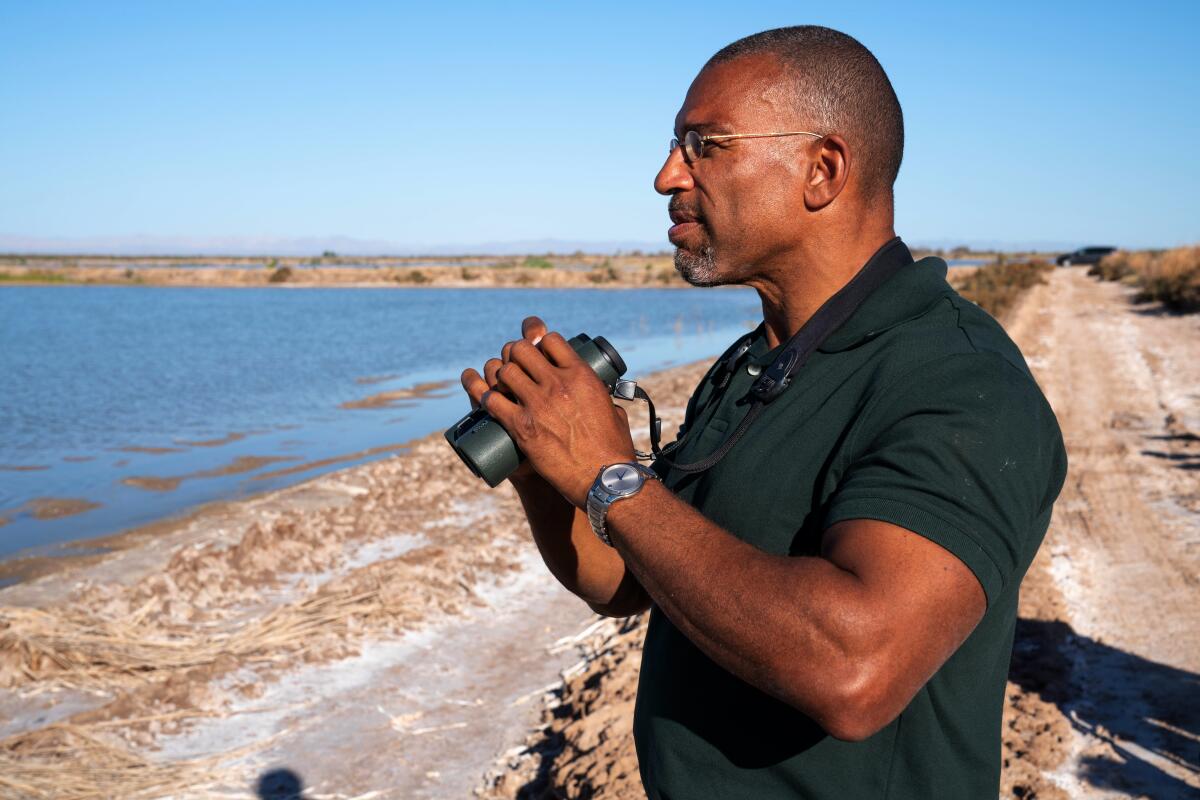 A man stands, holding binoculars, looking toward a body of water