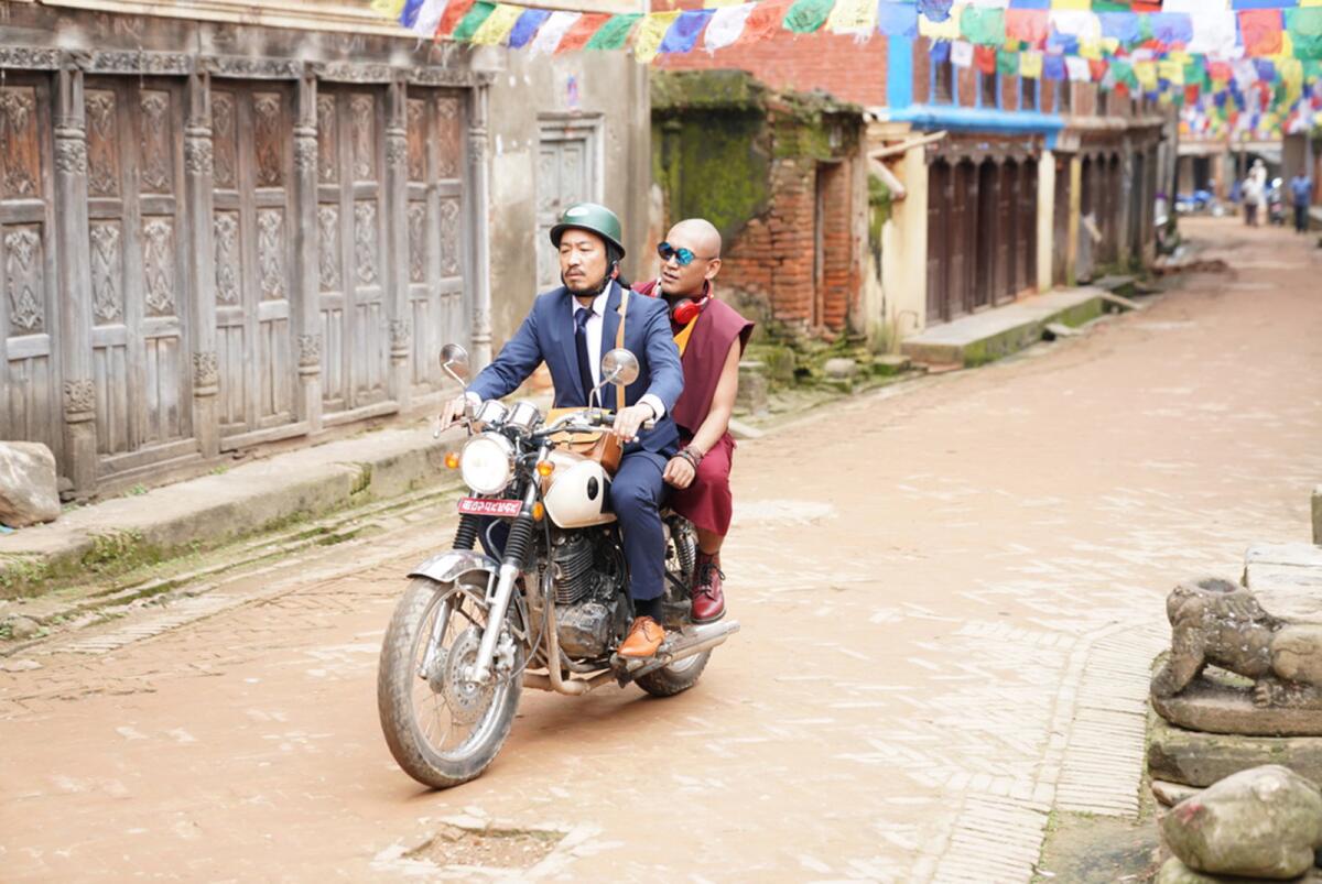 A man on a suit and a monk ride a motorbike.