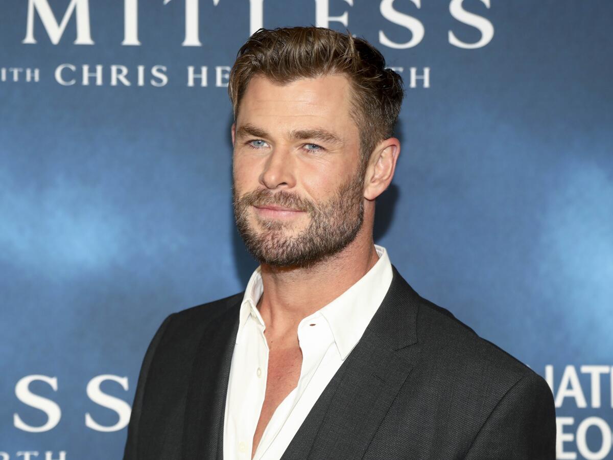 Chris Hemsworth in a black blazer and white suit shirt standing in front of a blue backdrop.