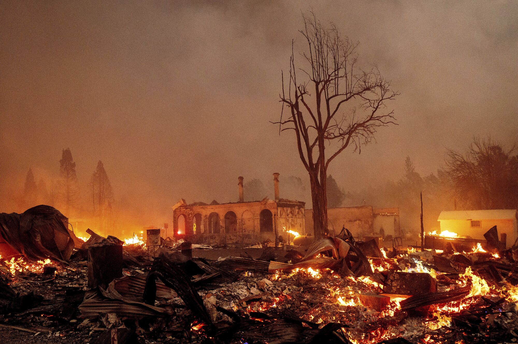 As dense smoke hangs over buildings, embers and flames cover the ground.