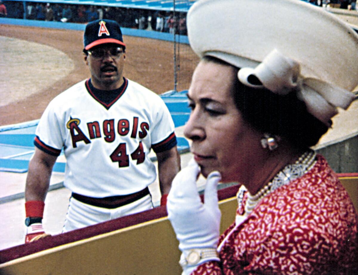 A baseball player in an Angels uniform stares at a woman in a large hat, white gloves and a red dress