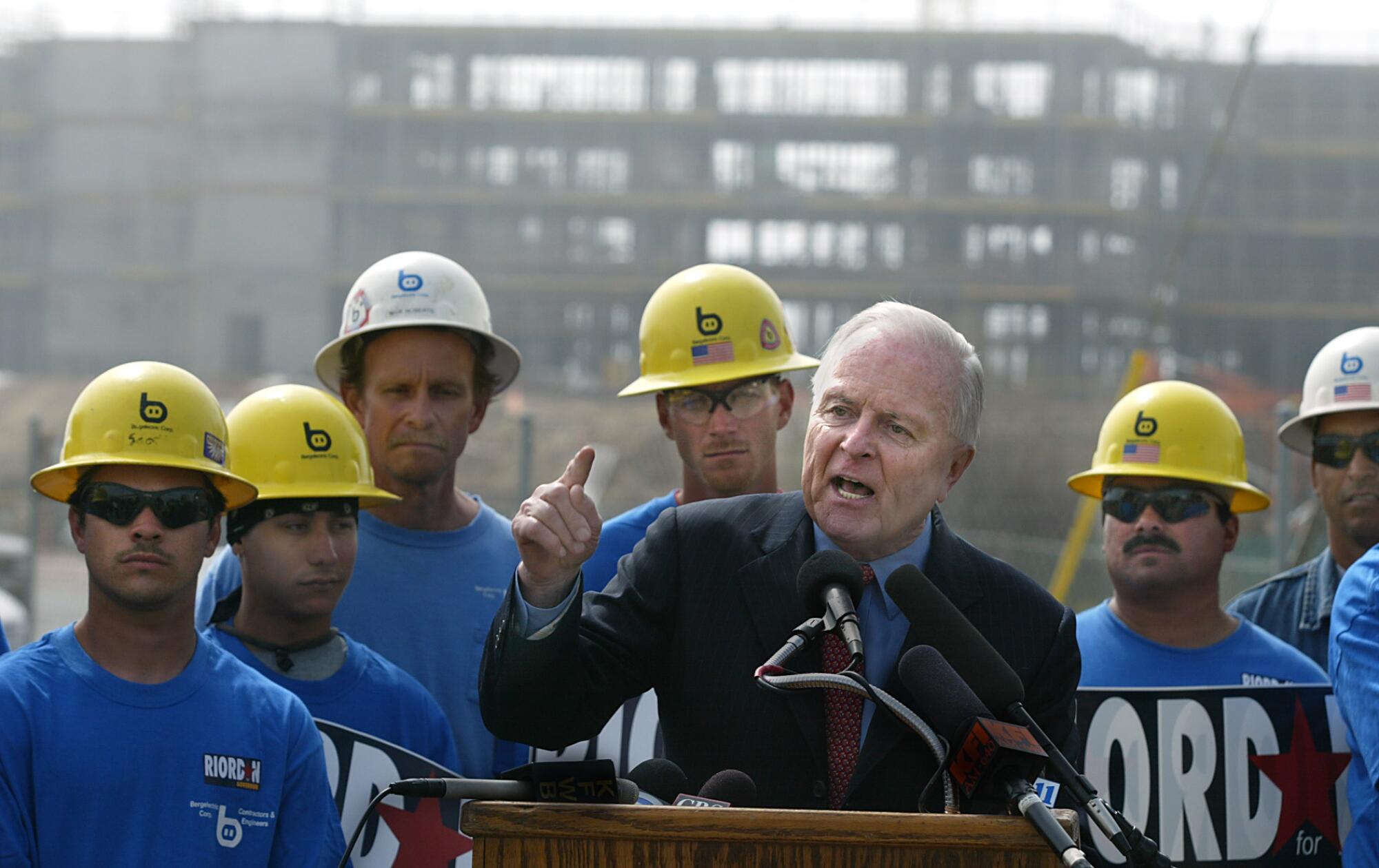 Richard Riordan speaks from a lectern with hardhats and the framework of a building under construction behind him