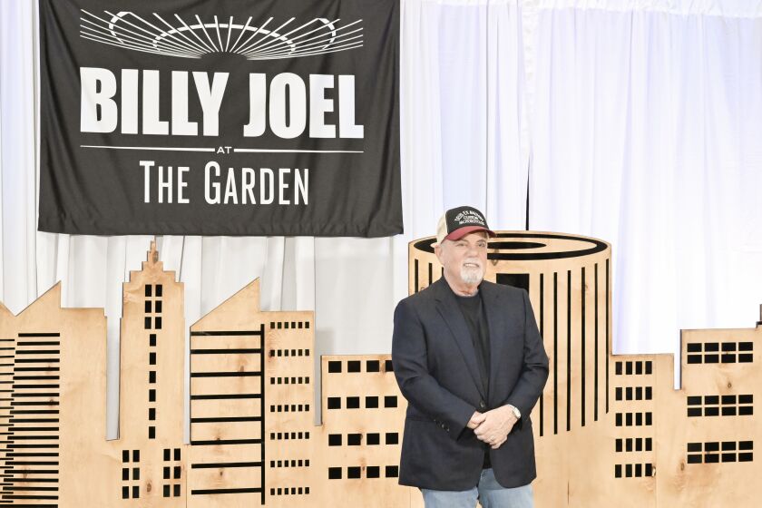 Billy Joel in a black shirt and blazer, jeans and a hat posing infant of a cloth backdrop