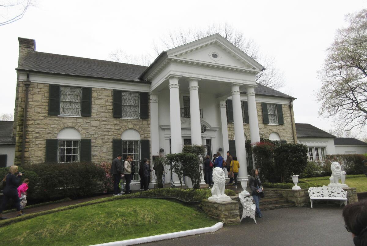 People stand in front of a mansion with a columned porch and statues flanking the steps in front
