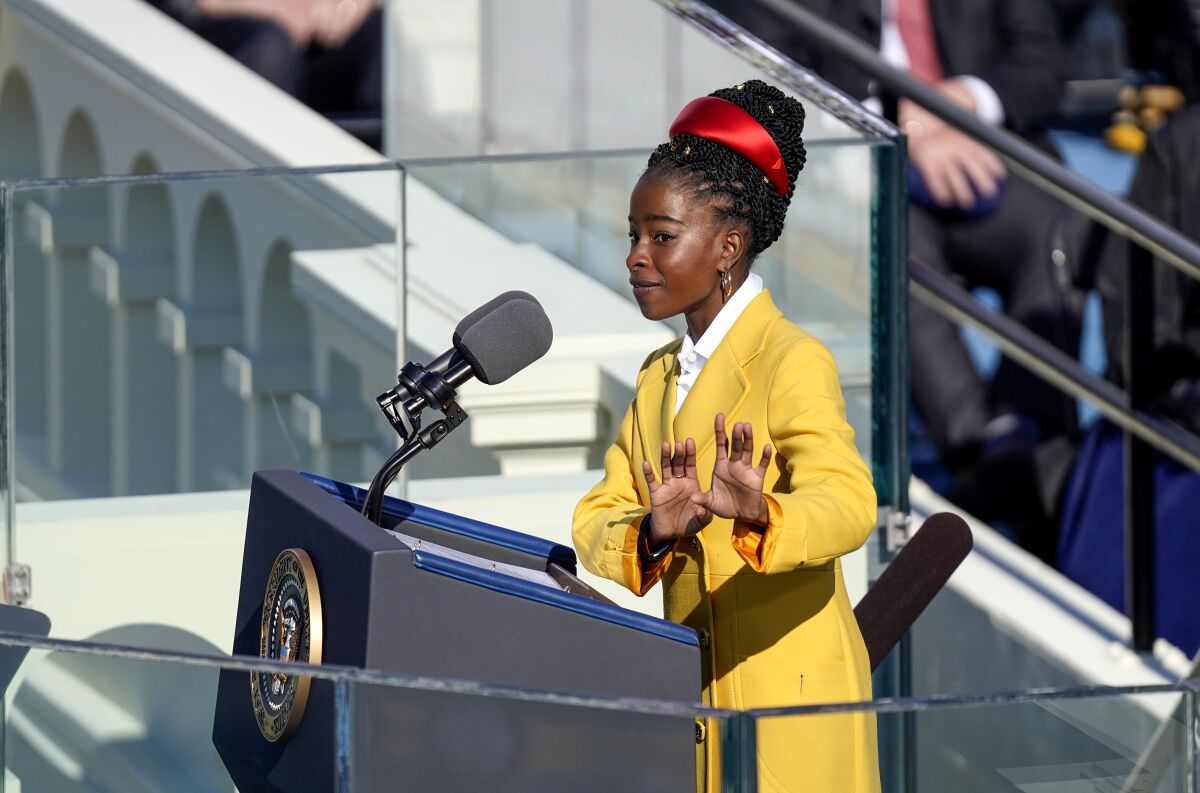 A young woman in a yellow coat gestures as she speaks before a lectern 