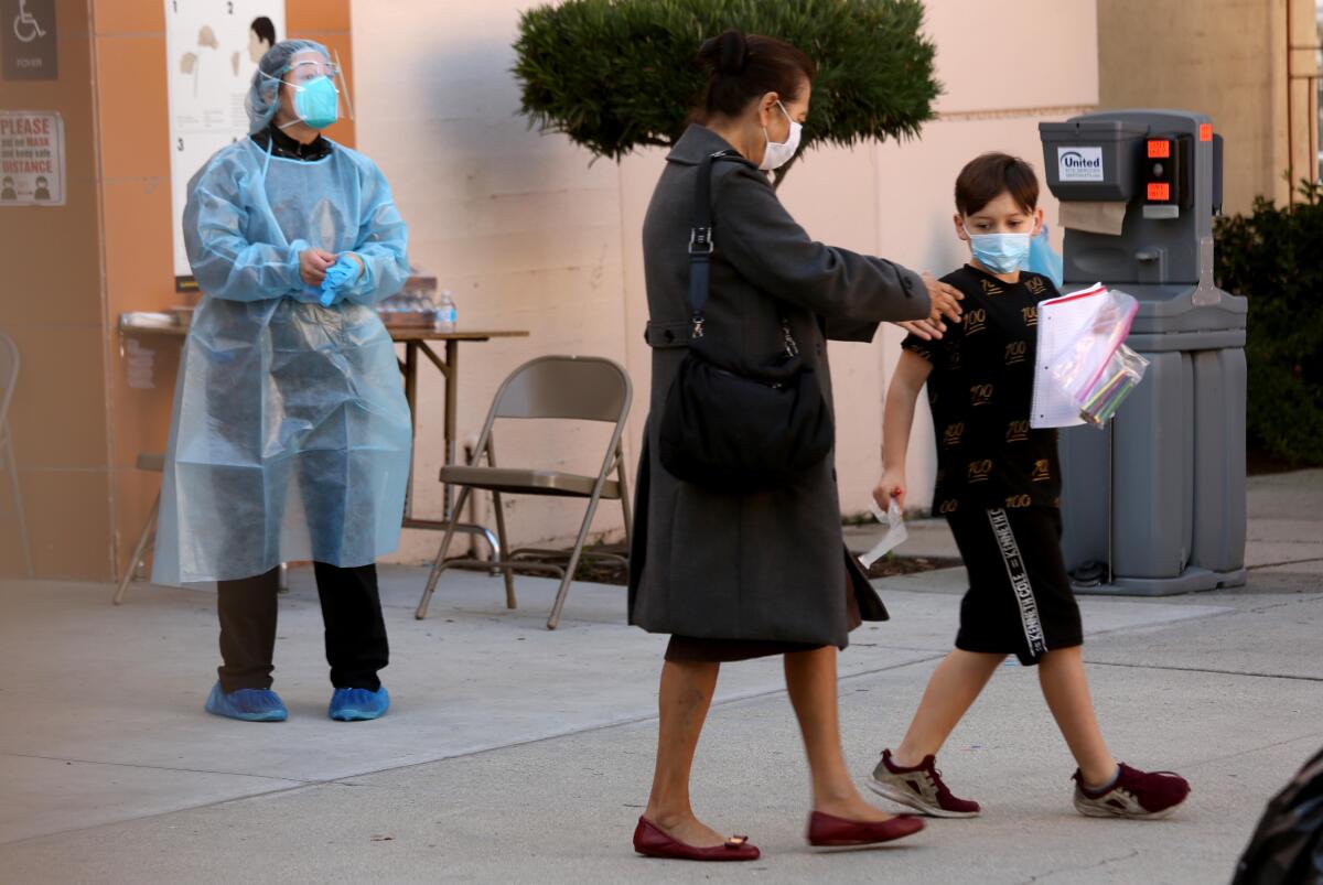 A woman walks next to a boy with a notebook and plastic bag in his hands as a worker in protective equipment looks on