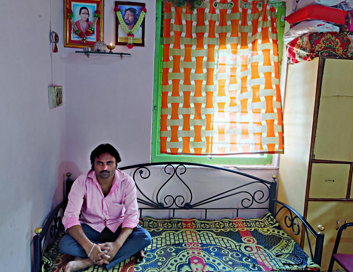A man sits on a daybed covered with a colorful bedspread under two photos and a lighted candle.